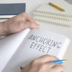 A person writing the words 'Anchoring Effect' in a lined notebook with a blue pen to define the cognitive bias.