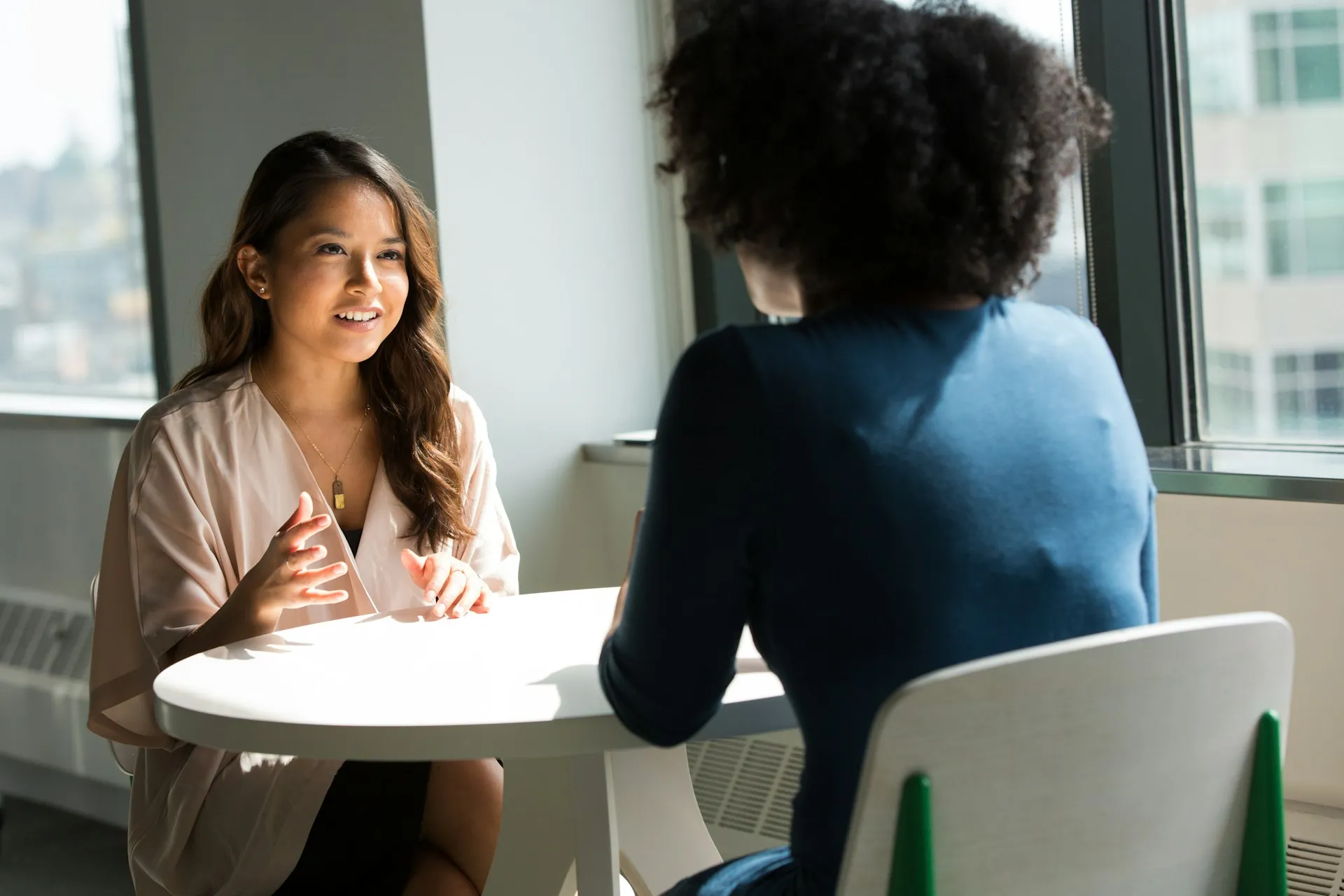 Two women sitting across a small table in conversation near a bright window