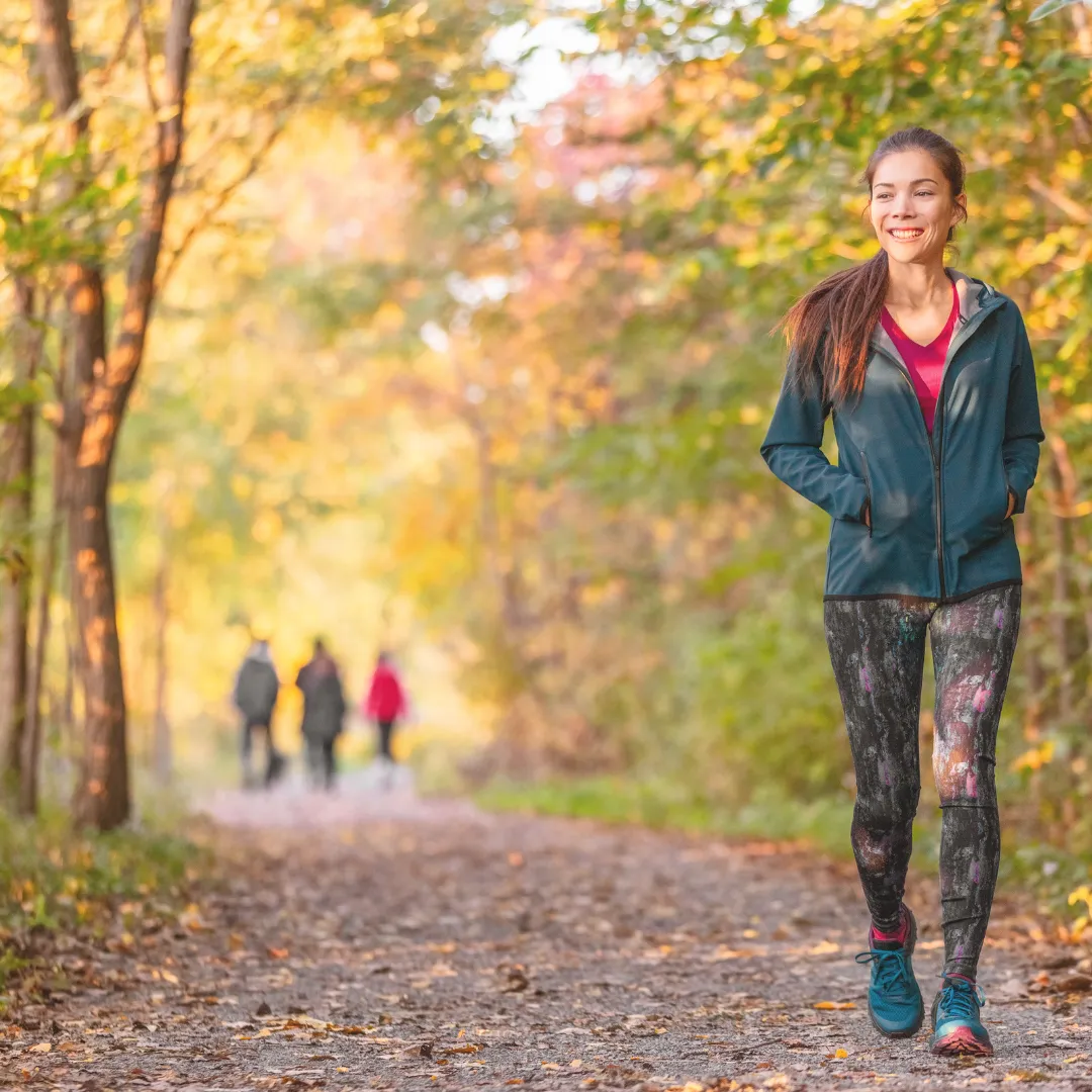 A person smiling while walking on a path through a forest with colorful autumn leaves, getting natural light to help with seasonal mood shifts.
