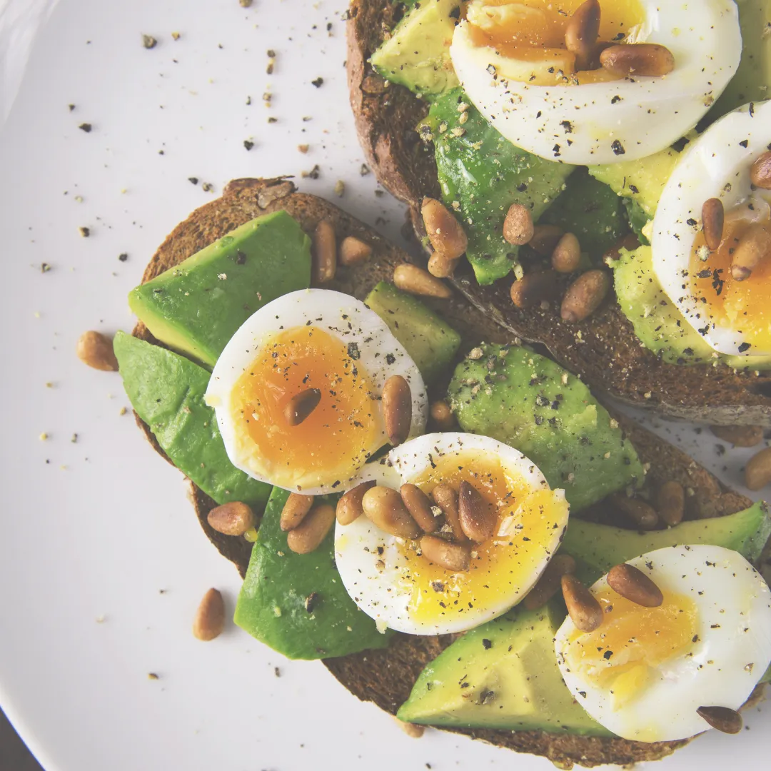 Close-up of two slices of rye toast topped with sliced avocado, soft-boiled eggs, and a sprinkle of black pepper and pine nuts.