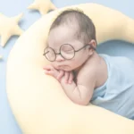 Newborn baby wearing glasses, peacefully sleeping on a moon-shaped cushion with stars in the background