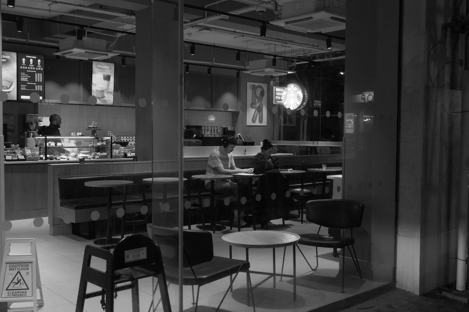 Two people working at a table inside a café seen through the window at night