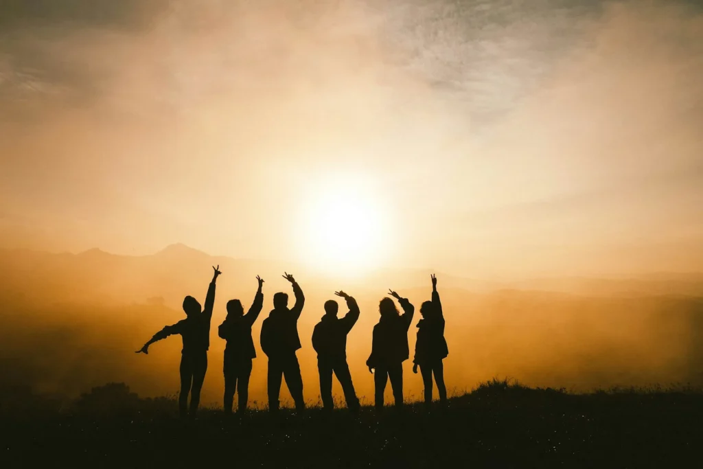 Six people silhouetted against a golden sunrise raising their arms on a hilltop