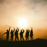 Six people silhouetted against a golden sunrise raising their arms on a hilltop