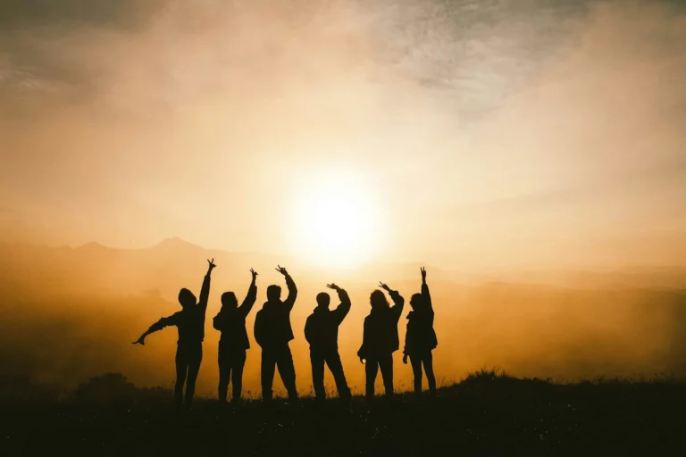 Six people silhouetted against a golden sunrise raising their arms on a hilltop