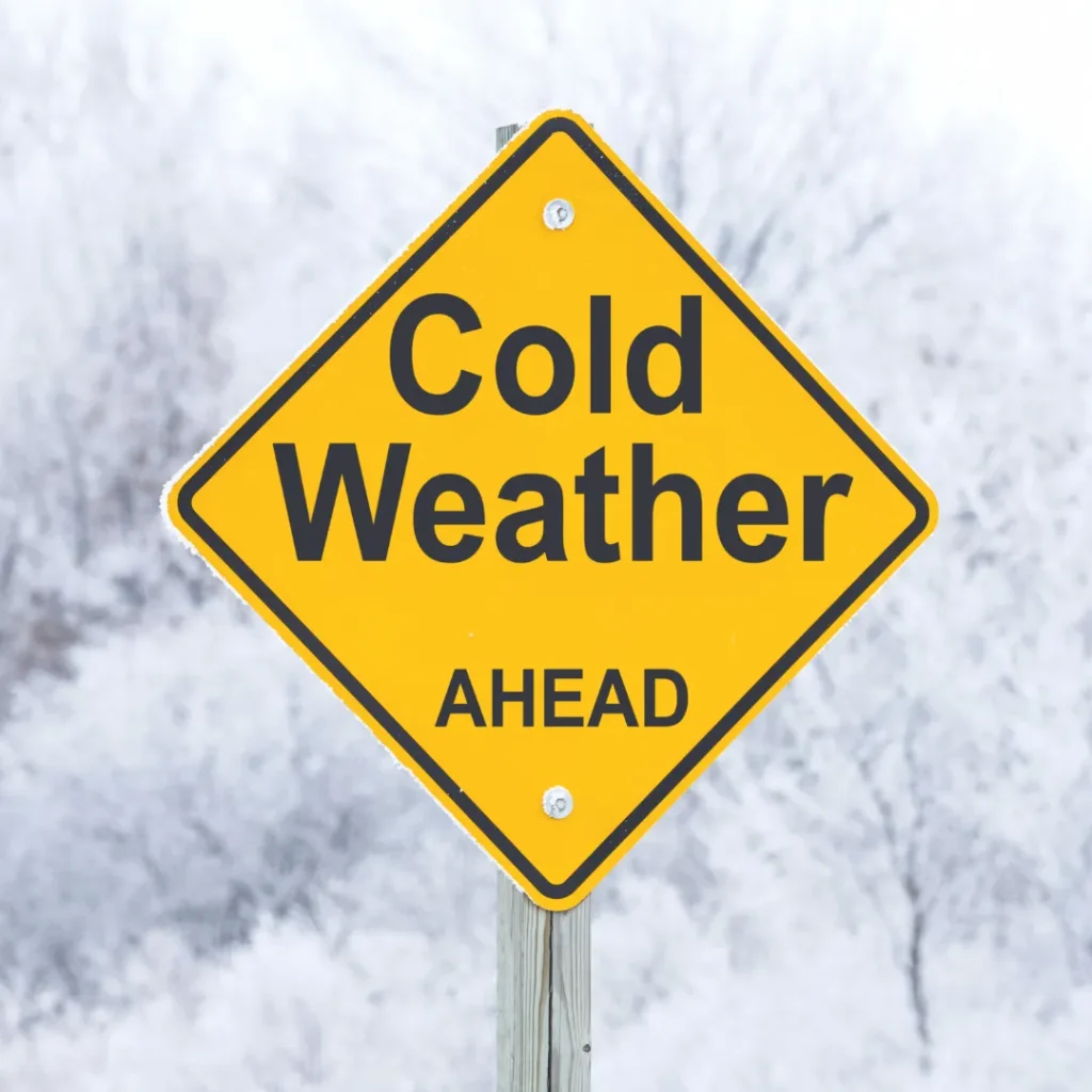 A yellow diamond road sign reading "Cold Weather Ahead" against a blurry snowy background, symbolizing the need to prepare the body's energy system for winter.