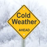 A yellow diamond road sign reading "Cold Weather Ahead" against a blurry snowy background, symbolizing the need to prepare the body's energy system for winter.