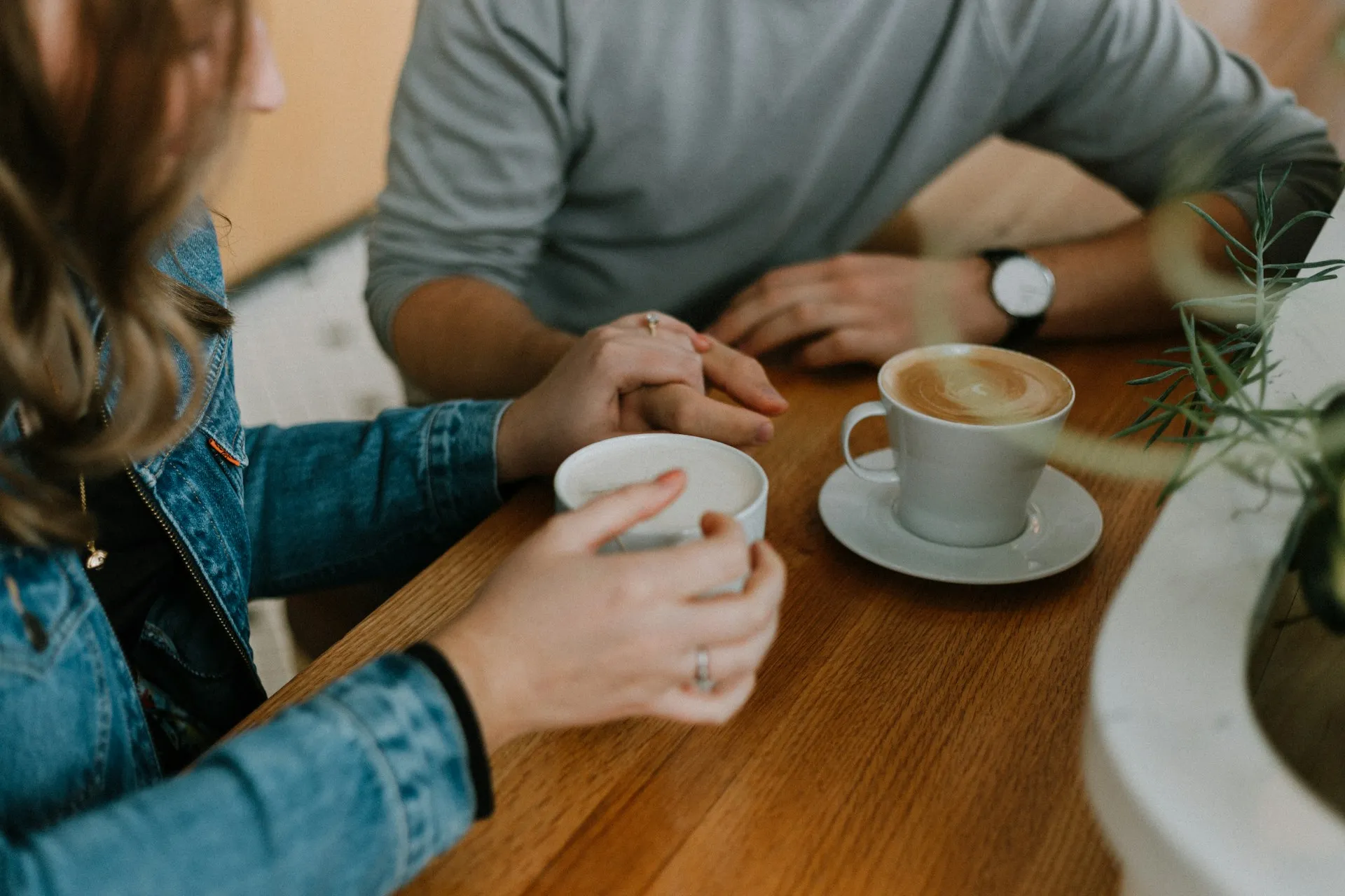 Two people sitting across a wooden table with coffee cups, hands resting near each other