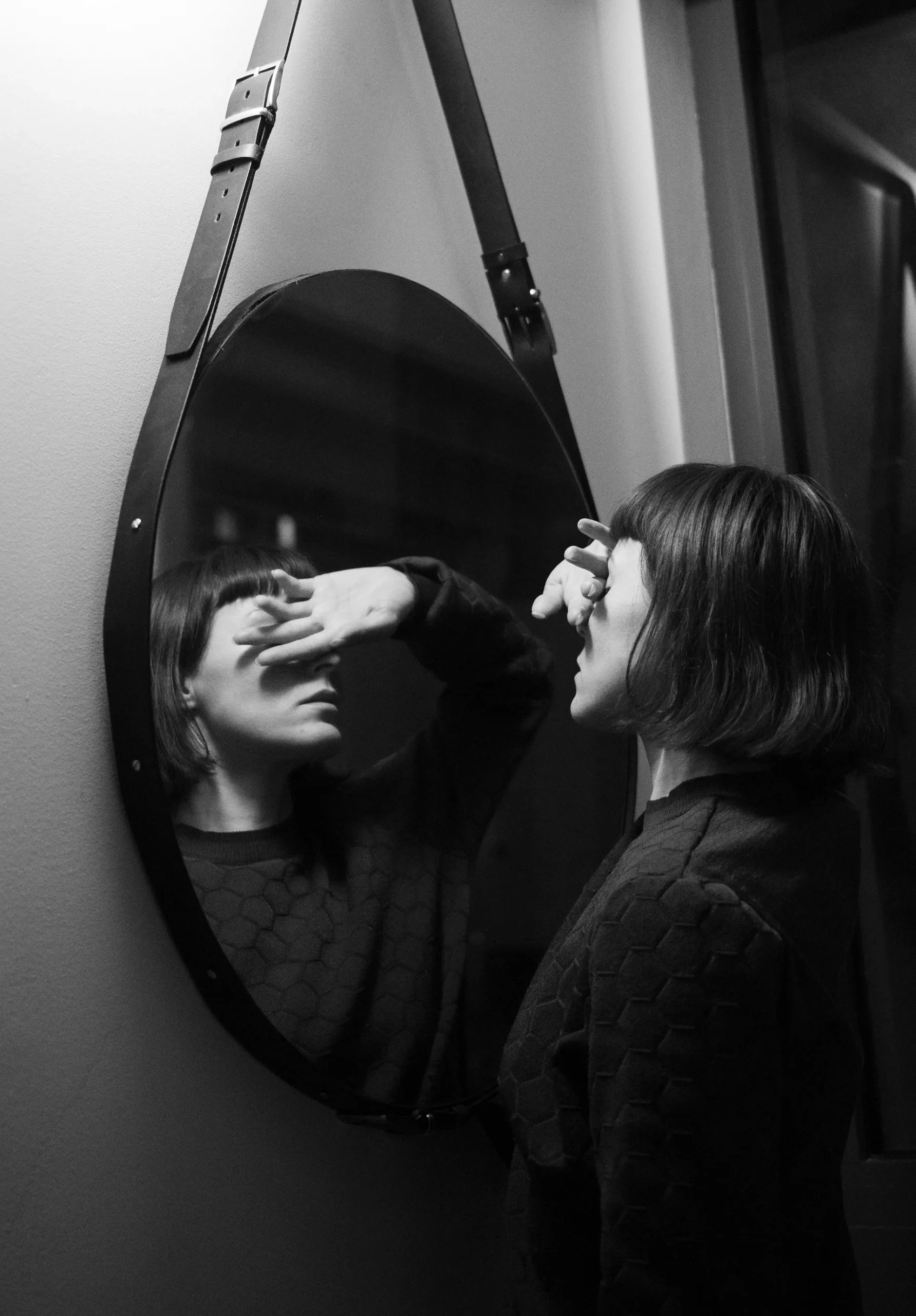 Black and white photo of a woman standing at a round wall mirror covering her eyes with one hand