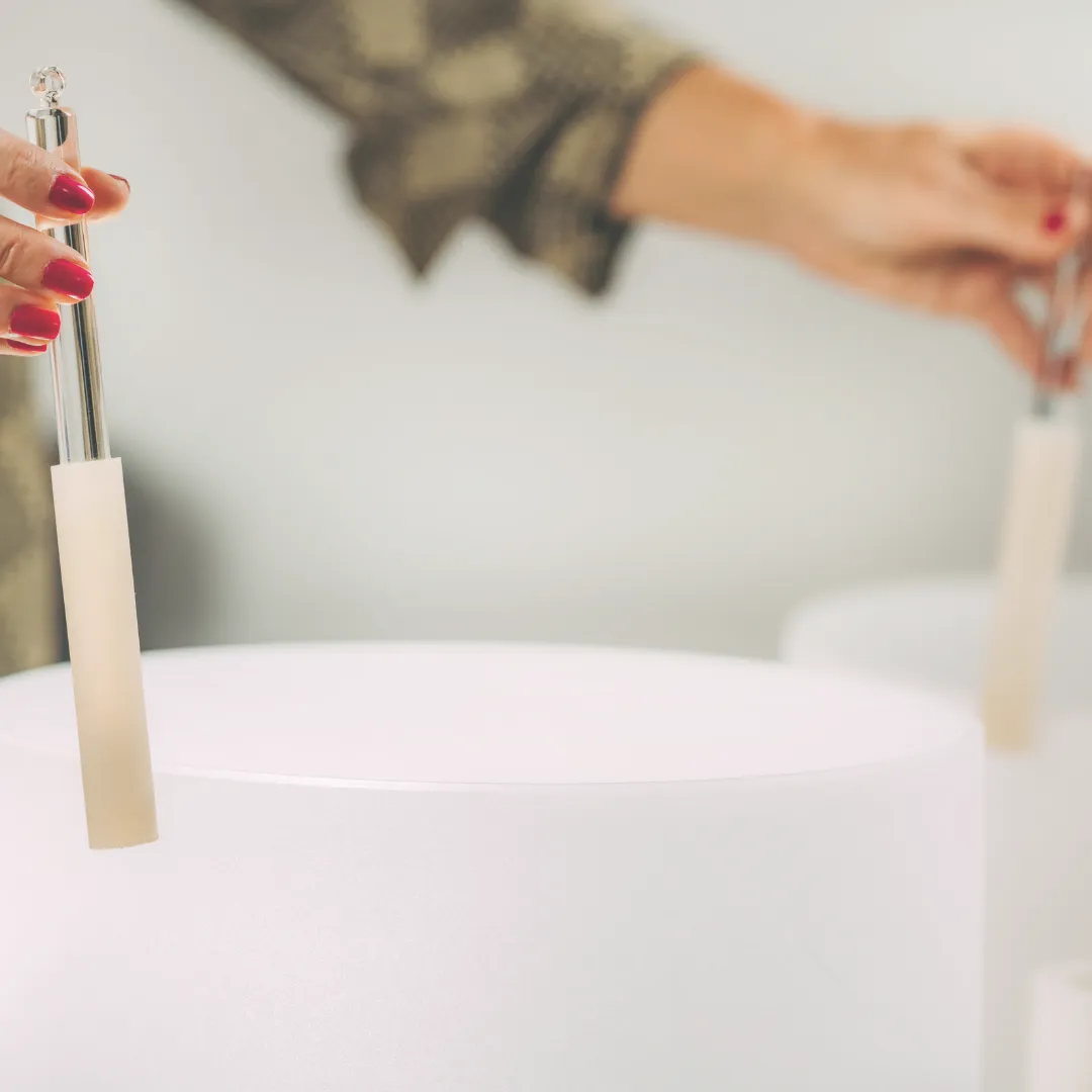 A sound healing practitioner holds two mallets over large white crystal singing bowls during a biofield therapy session.