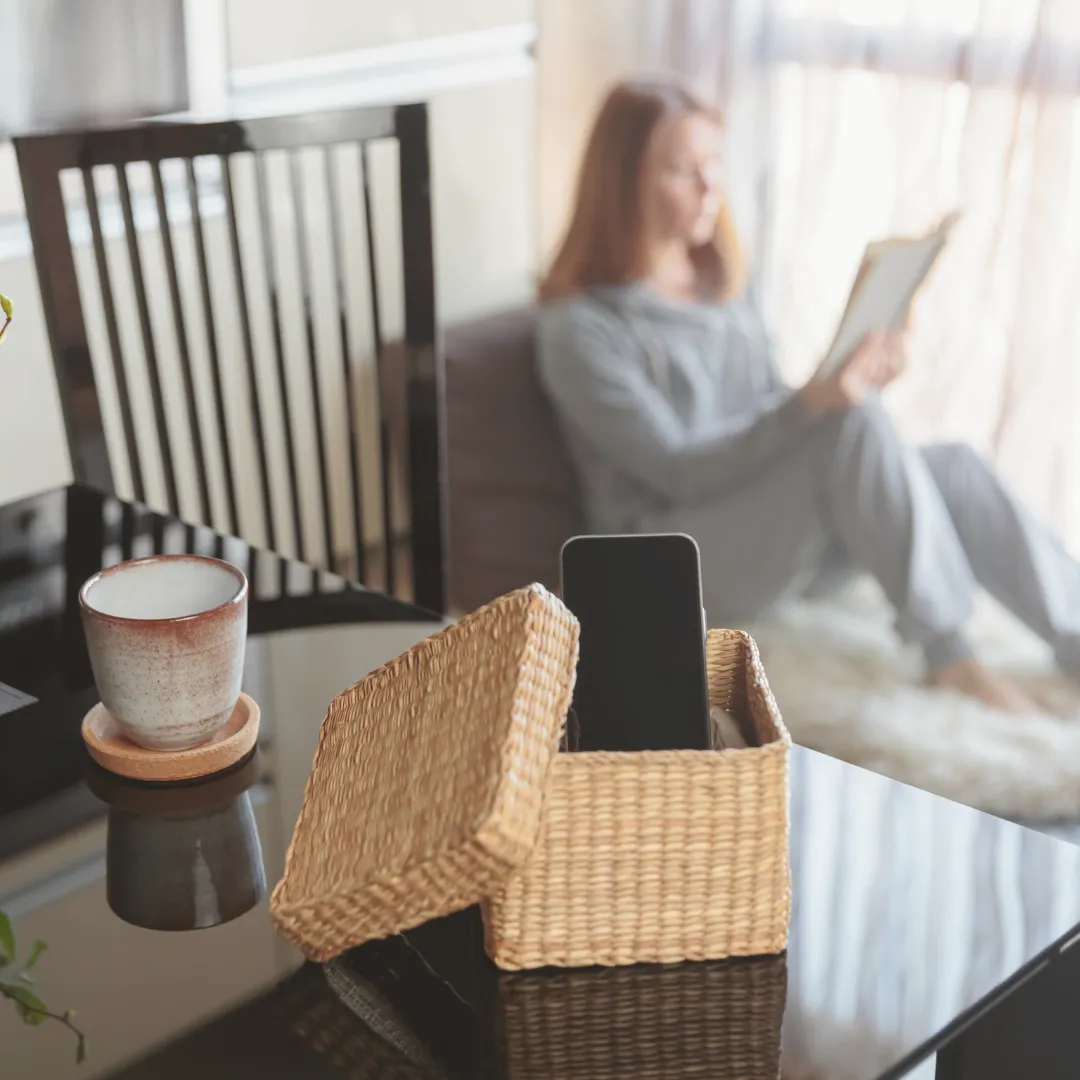 A smartphone placed inside a wicker box, with a person relaxing and reading a book in the background, symbolizing a digital detox for wellness.