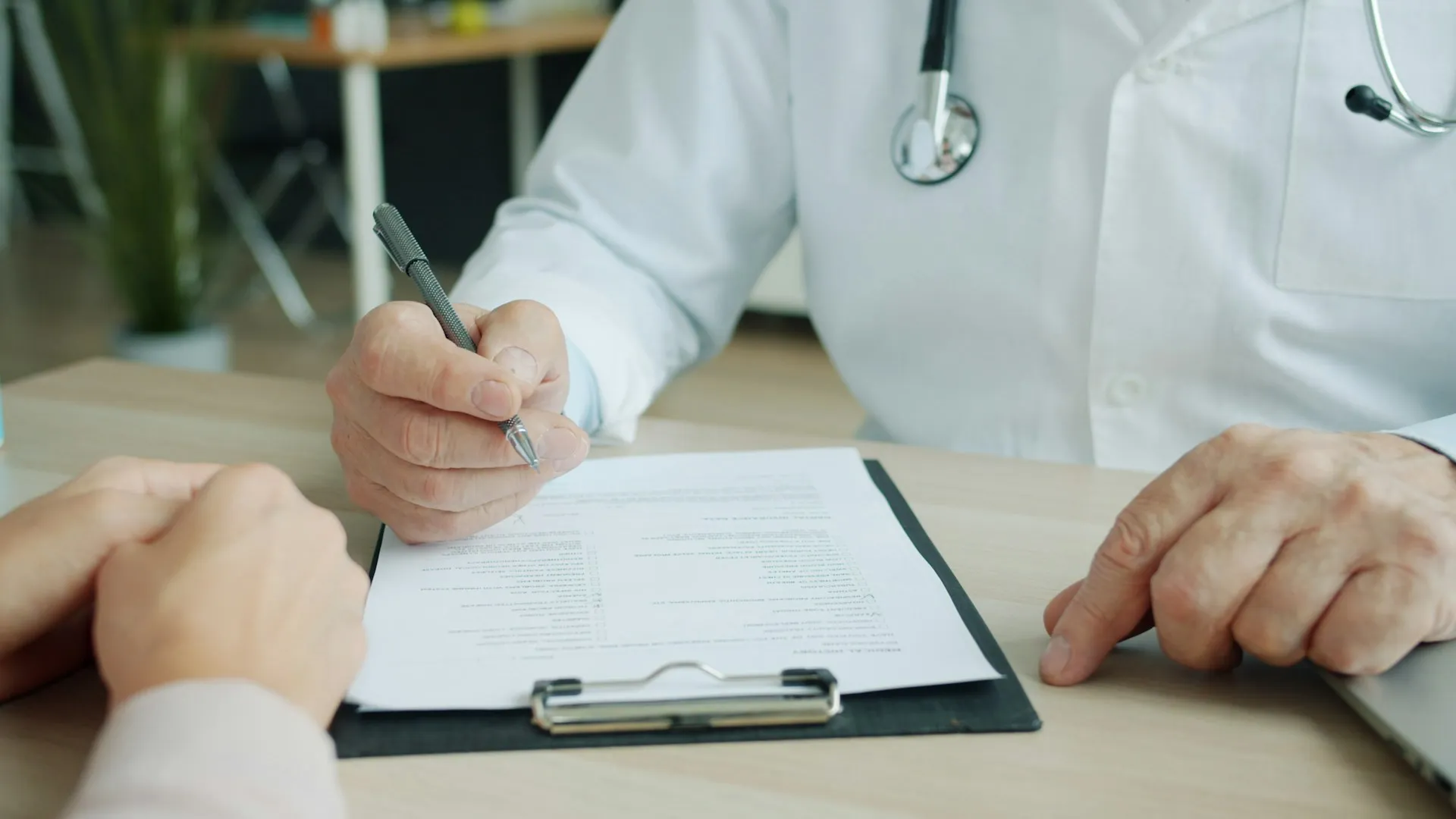 Doctor writing on a clipboard during a consultation while a patient sits across the desk