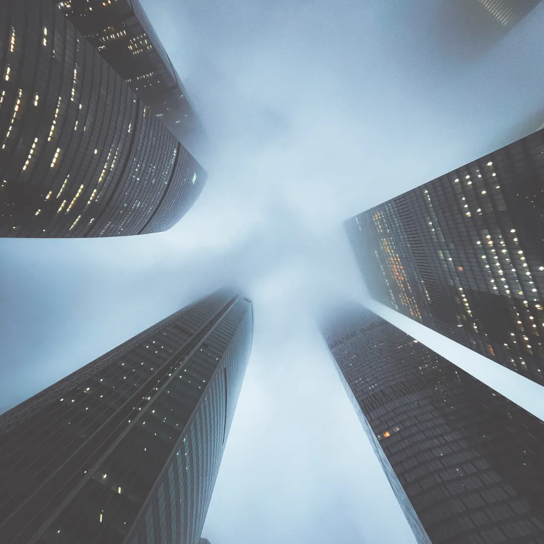 A low-angle view looking up at modern skyscrapers shrouded in fog, symbolizing the feeling of being overwhelmed by EMFs and energetic congestion.