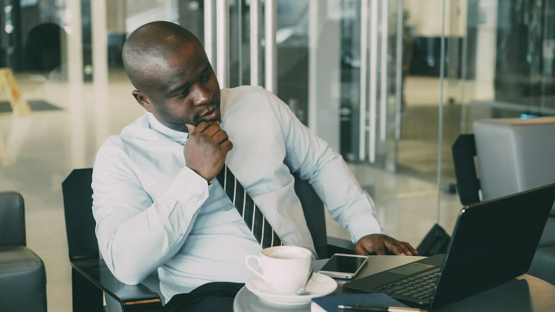 Man in a shirt and tie working thoughtfully at a laptop with coffee beside him