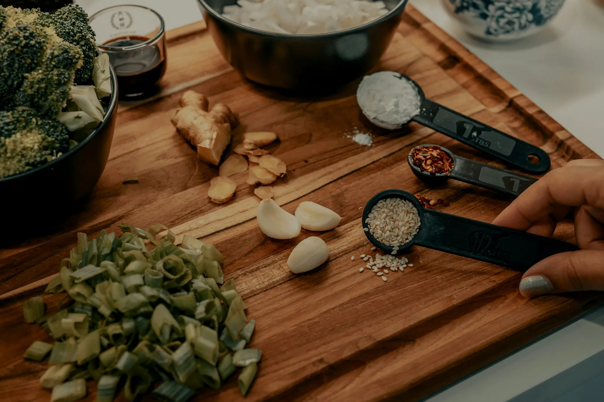 Chopped scallions, garlic, ginger, and broccoli on a wooden cutting board with measuring spoons