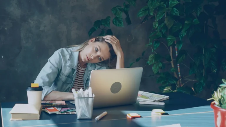 Woman in a blazer at a white desk with both hands pressed to her temples in front of a laptop