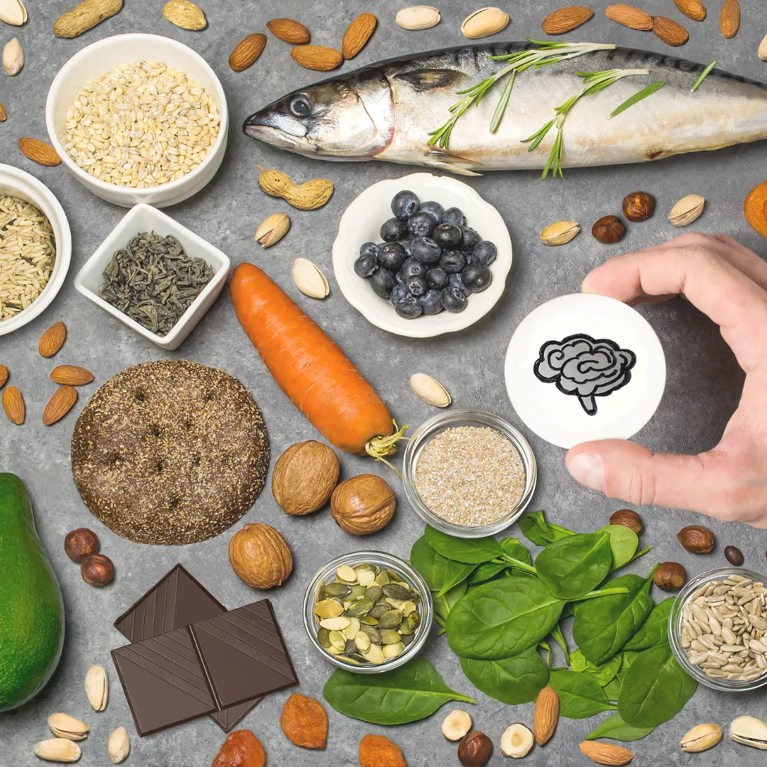 An overhead view of brain-healthy foods like fish, nuts, and leafy greens, with a hand holding a symbol of a brain, illustrating the gut-brain connection in functional medicine.