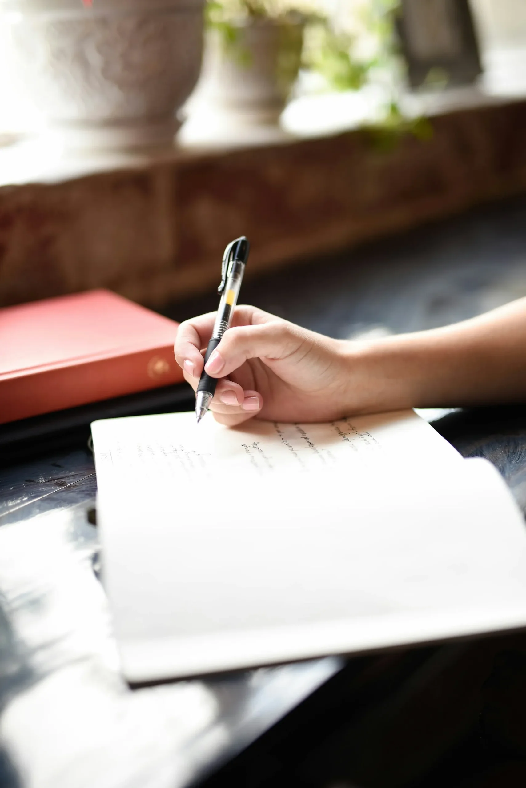 Hand writing in an open notebook on a table beside a red book and sunlit window