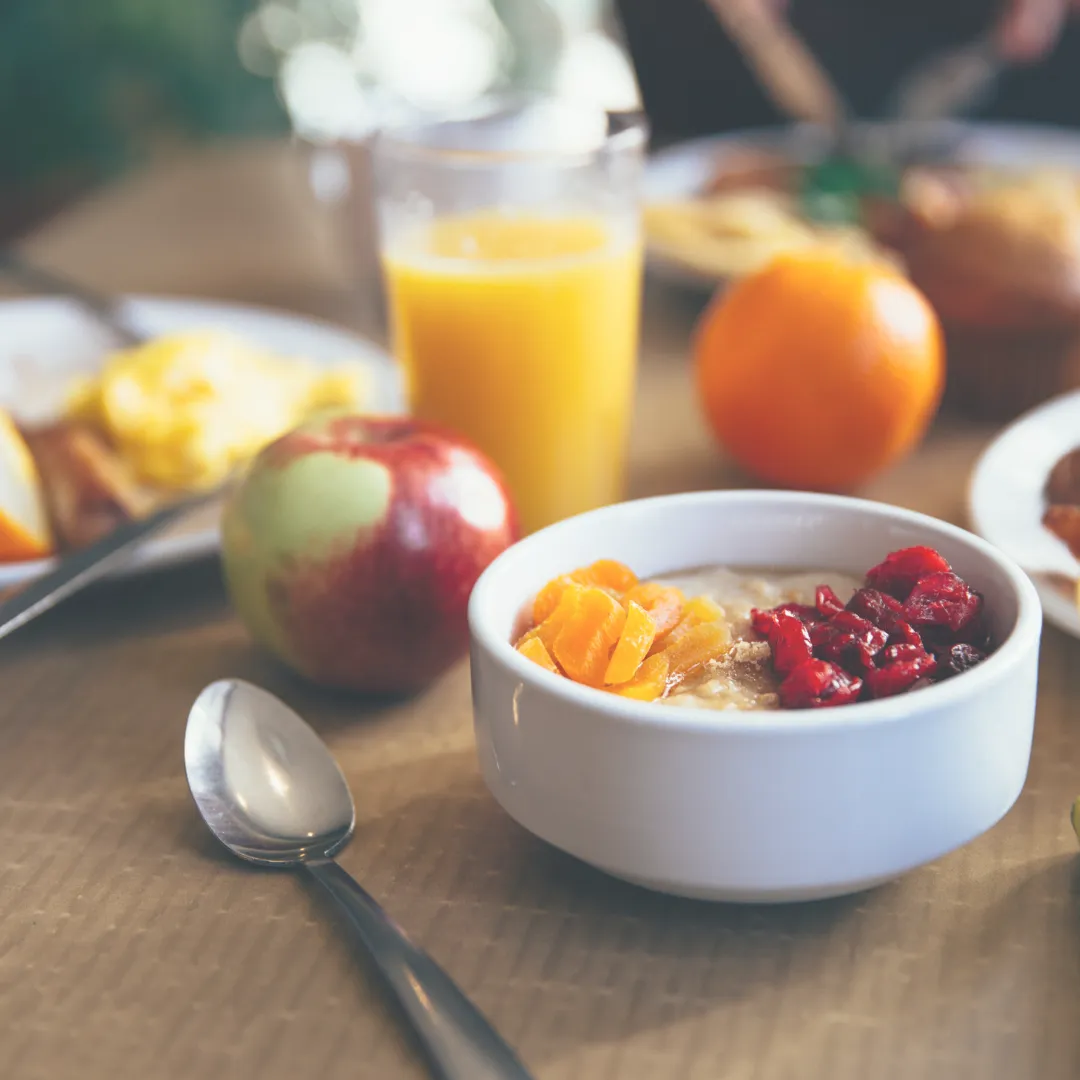 A white bowl of oatmeal topped with dried cranberries and apricots, next to a fresh apple and a glass of orange juice.