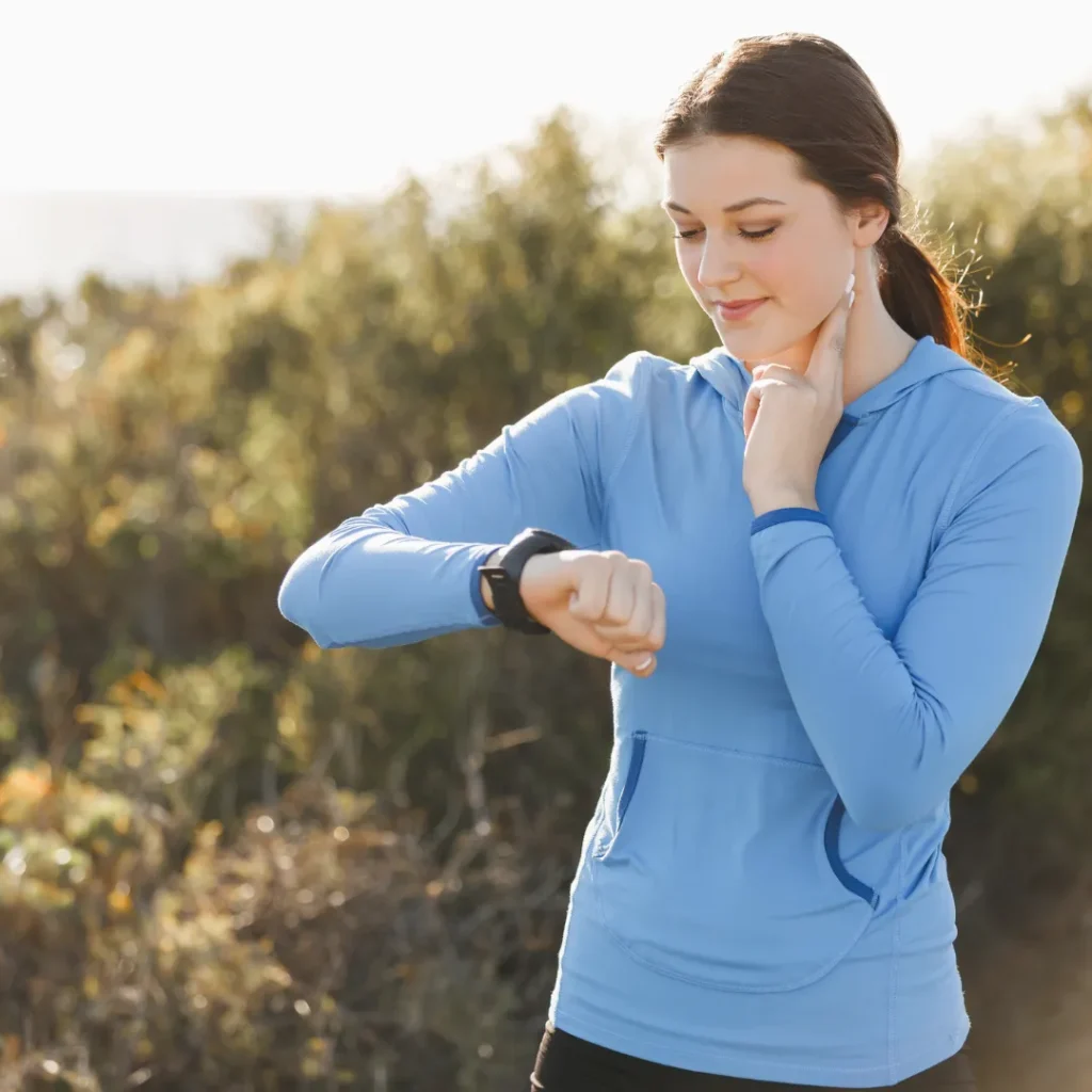 A female athlete in athletic wear checking her wrist-worn heart rate monitor while also manually checking her pulse, symbolizing daily HRV monitoring for training readiness.