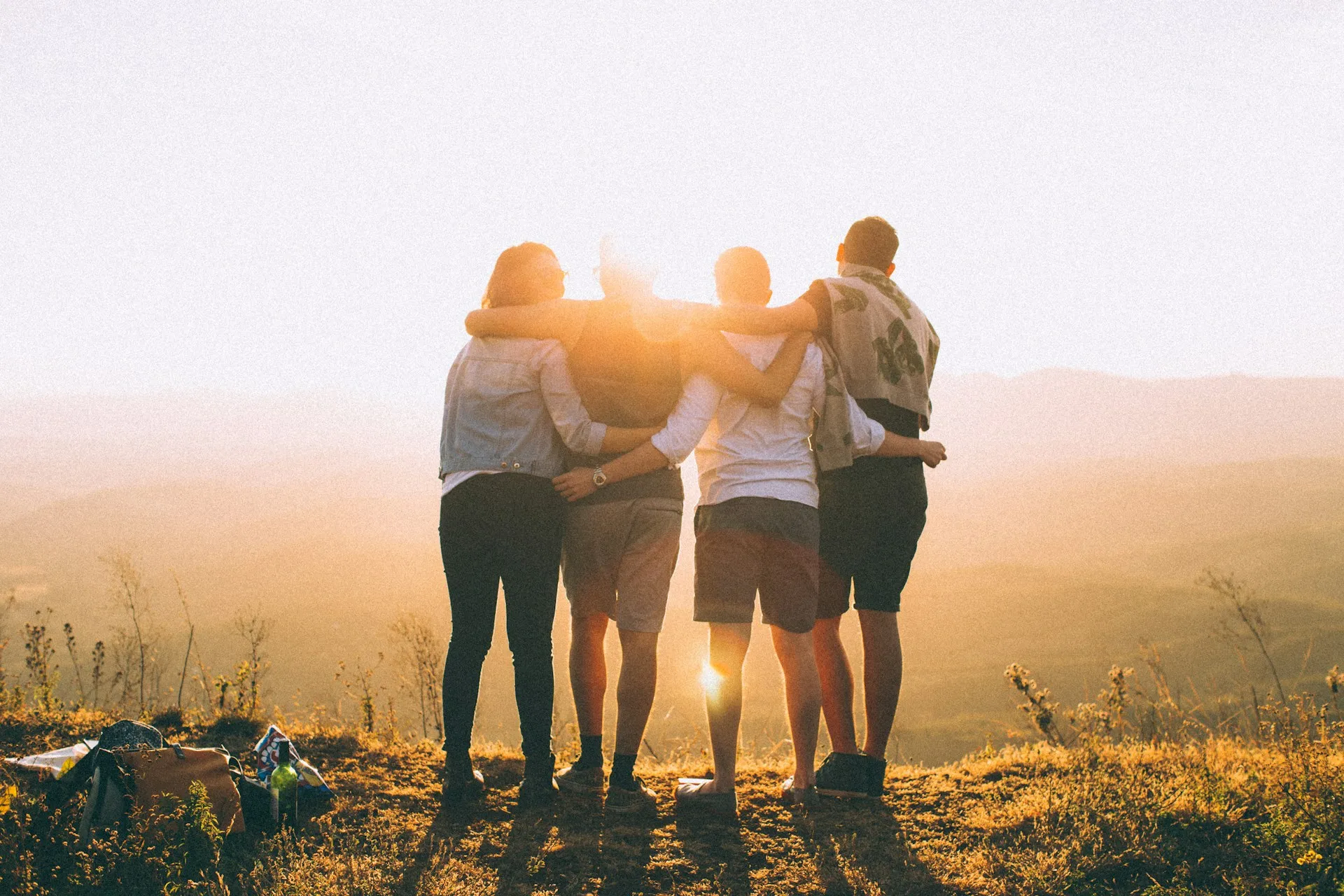 Four friends standing with arms around each other looking out over hills at sunset