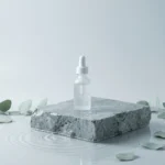 A clear serum dropper bottle on a wet grey stone block surrounded by green eucalyptus leaves in water, symbolizing holistic support for summer fatigue and allergies.