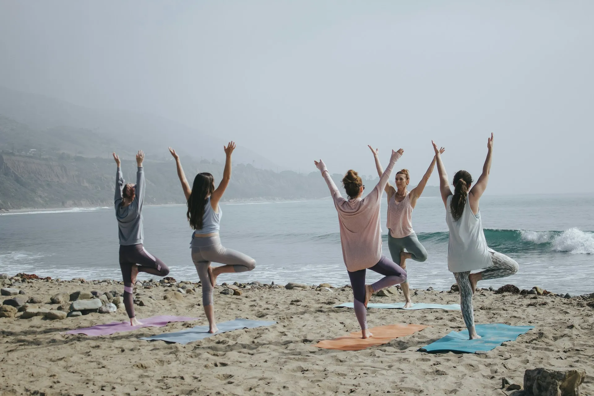 Five people practicing tree pose together on yoga mats on a foggy beach