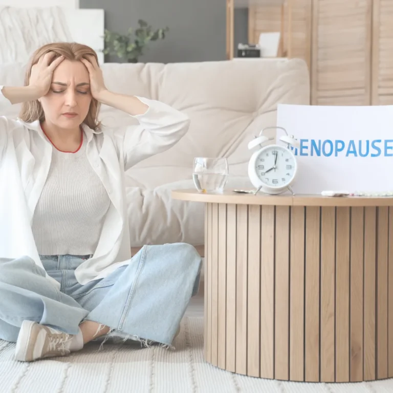 Woman experiencing menopausal symptoms like fatigue or headache, sitting near a menopause awareness sign.