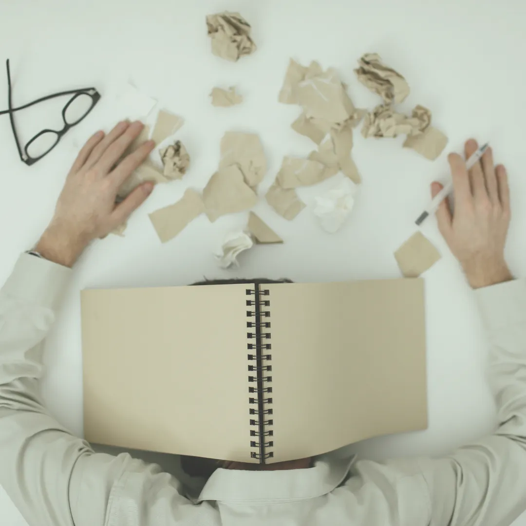 A man resting their head on a messy desk with a laptop and paperwork, showing the physical and mental exhaustion that reduces motivation for health behaviors.