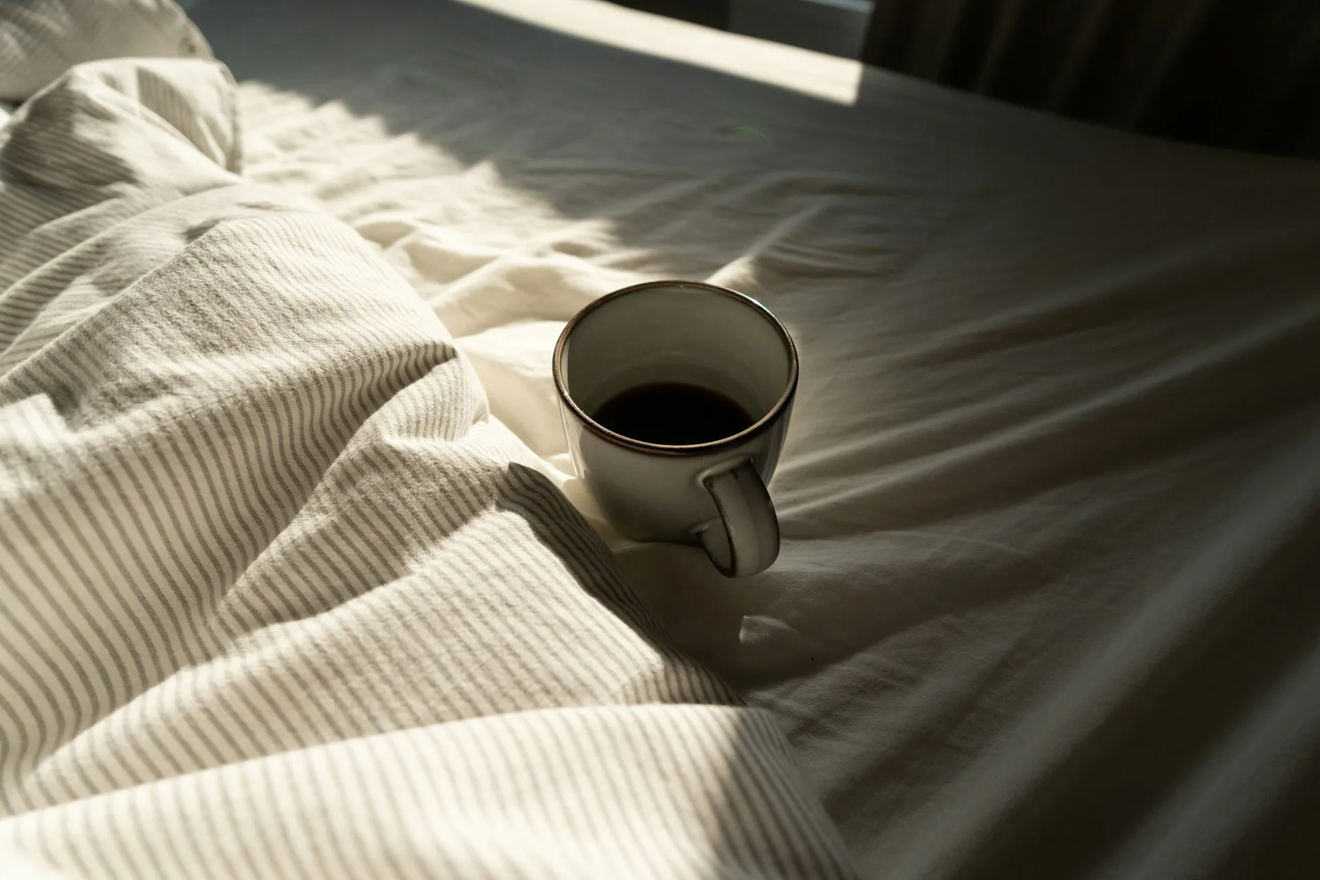 Mug of coffee on rumpled bed linens with morning sunlight falling across them