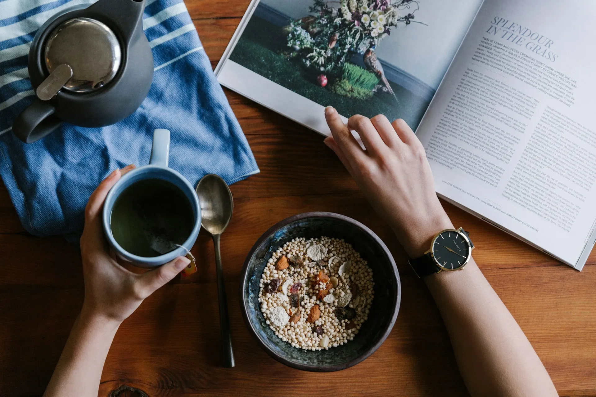 Overhead view of hands holding a mug of tea beside a breakfast bowl and open magazine