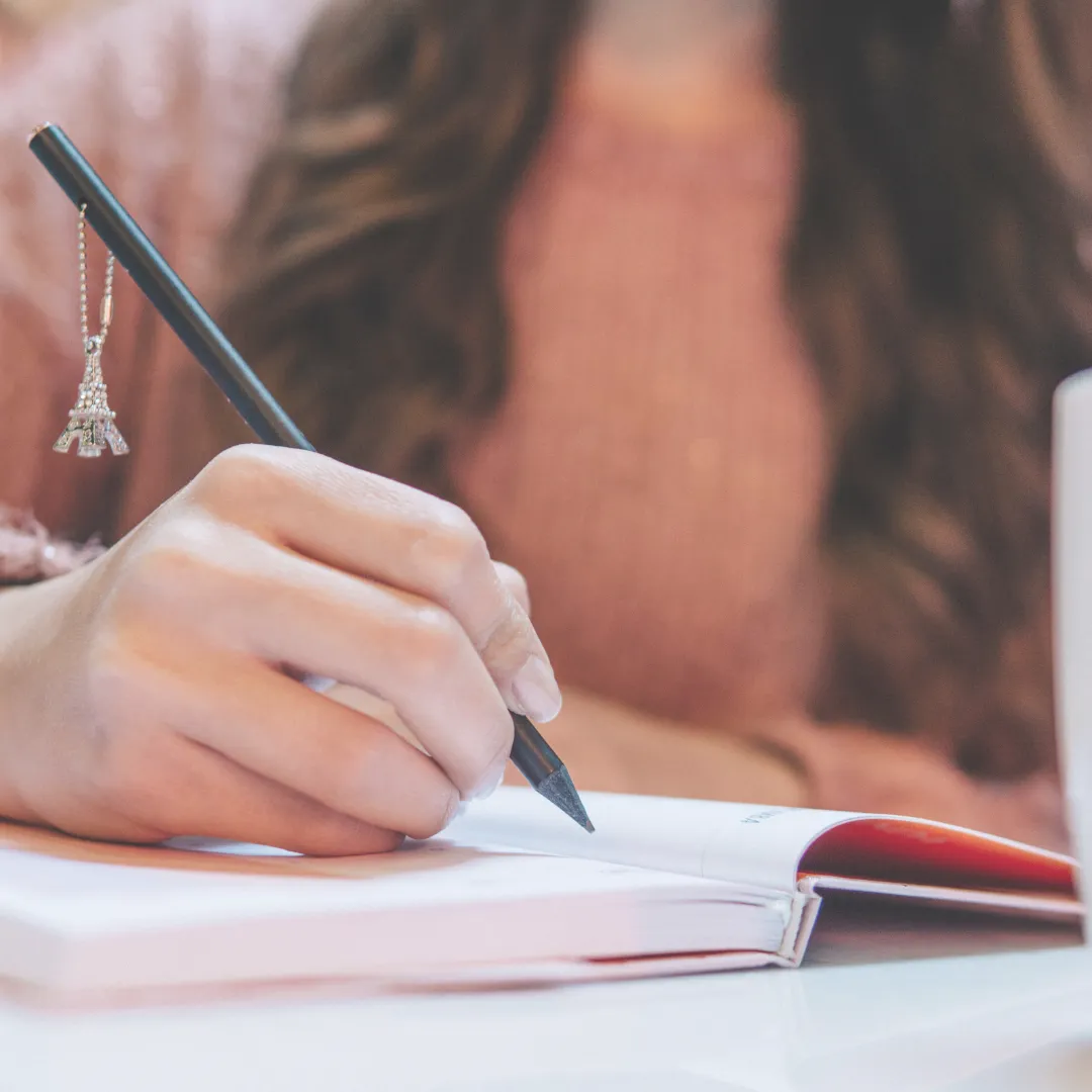 A close-up of a person's hands writing with a pencil in a notebook, symbolizing the re-authoring practice in narrative therapy.