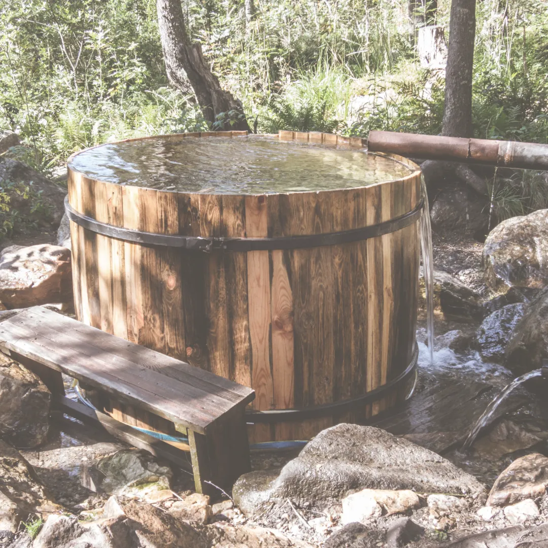 A rustic wooden tub set up as a cold plunge in a natural, rocky forest setting, with fresh water flowing into it.