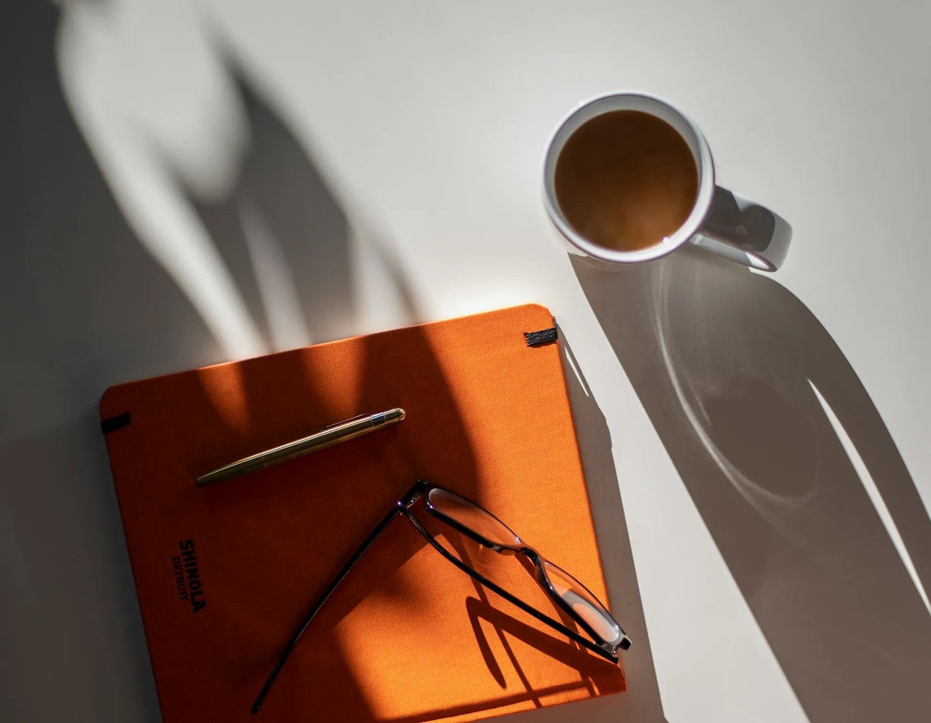 Orange notebook with a gold pen and reading glasses beside a mug of coffee in hard sunlight