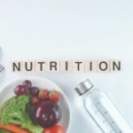 The word NUTRITION spelled out in wooden blocks, with a bowl of fresh fruits and vegetables and a bottle of water below it.