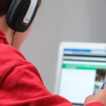 Student in a red hoodie wearing headphones while working at a laptop with a pen in hand