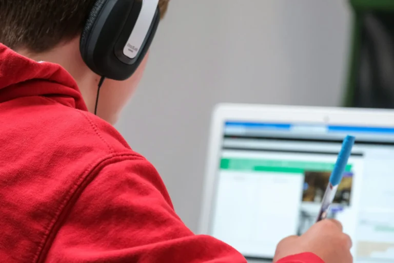 Student in a red hoodie wearing headphones while working at a laptop with a pen in hand