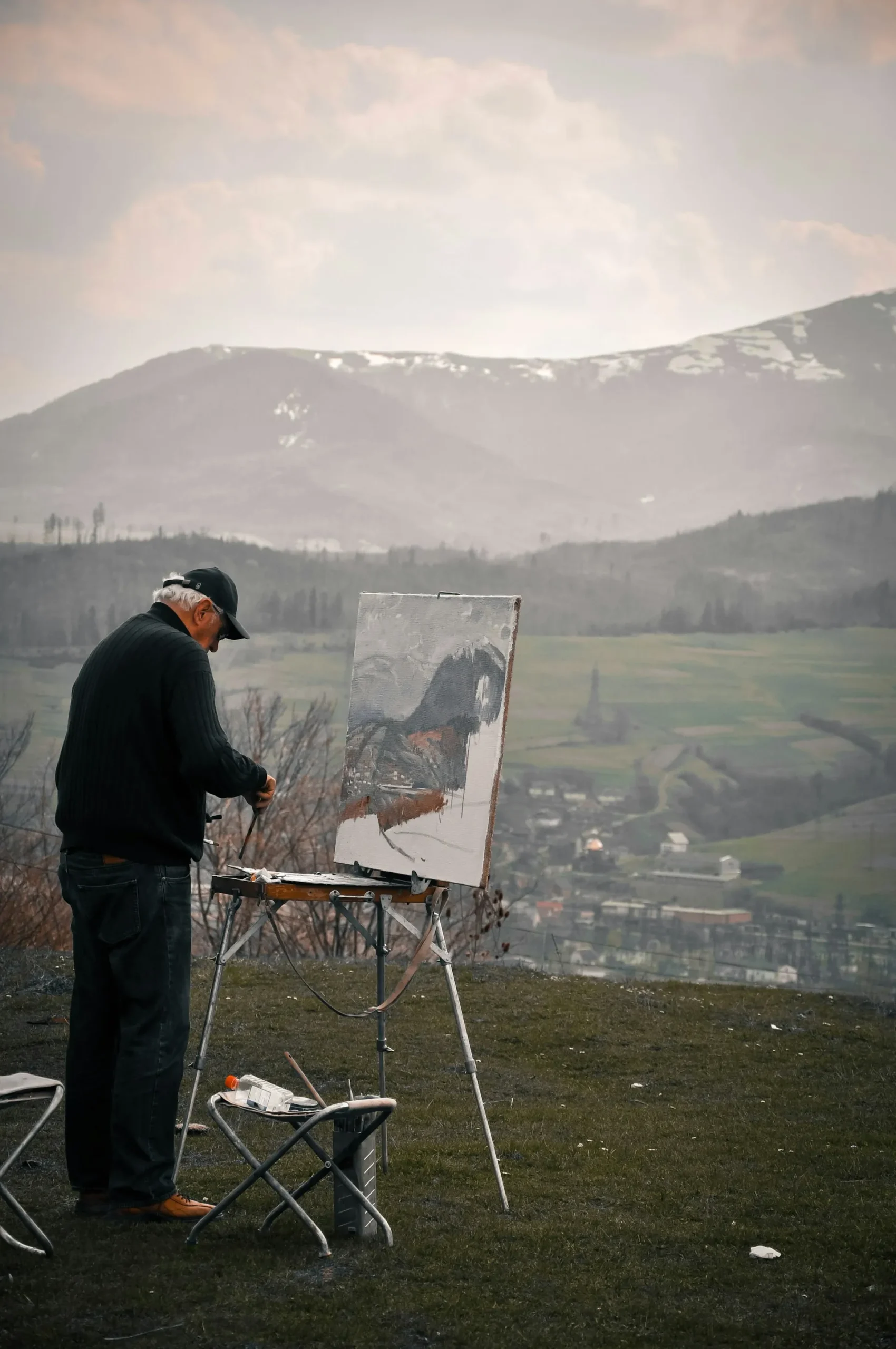 Older man painting at an easel on a hillside overlooking a valley and snow-topped mountains