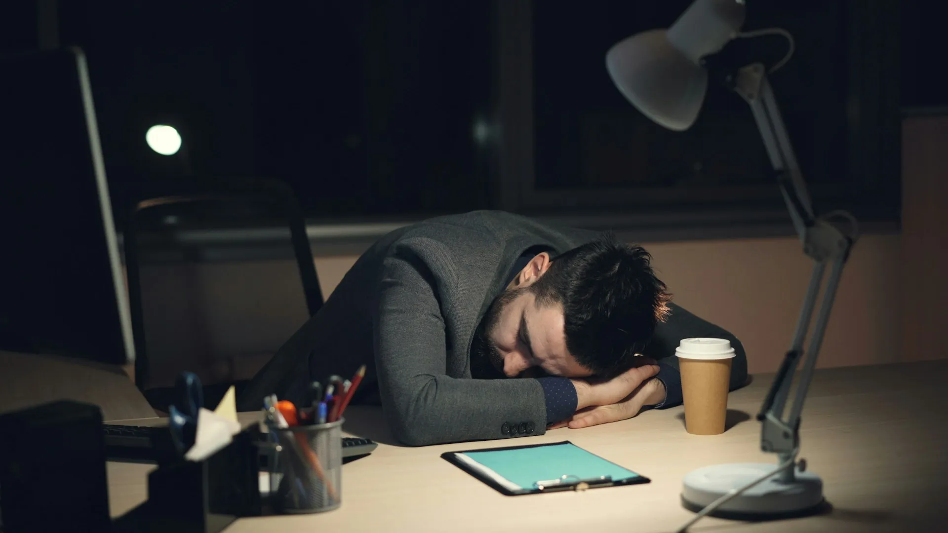 Man in a suit jacket resting his head on a desk beside a coffee cup in a dim office