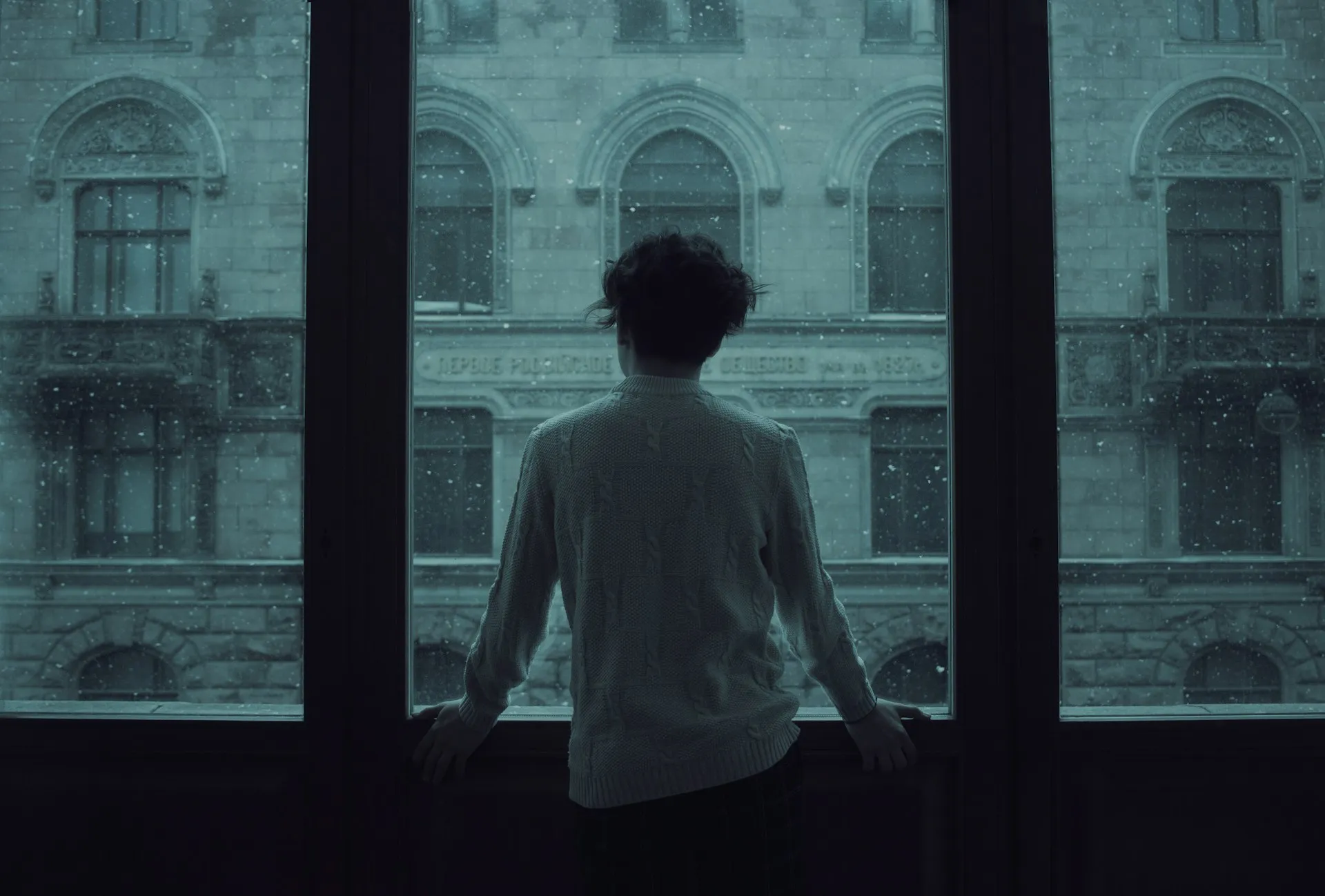 Person standing alone looking out a large window at a snowy building facade