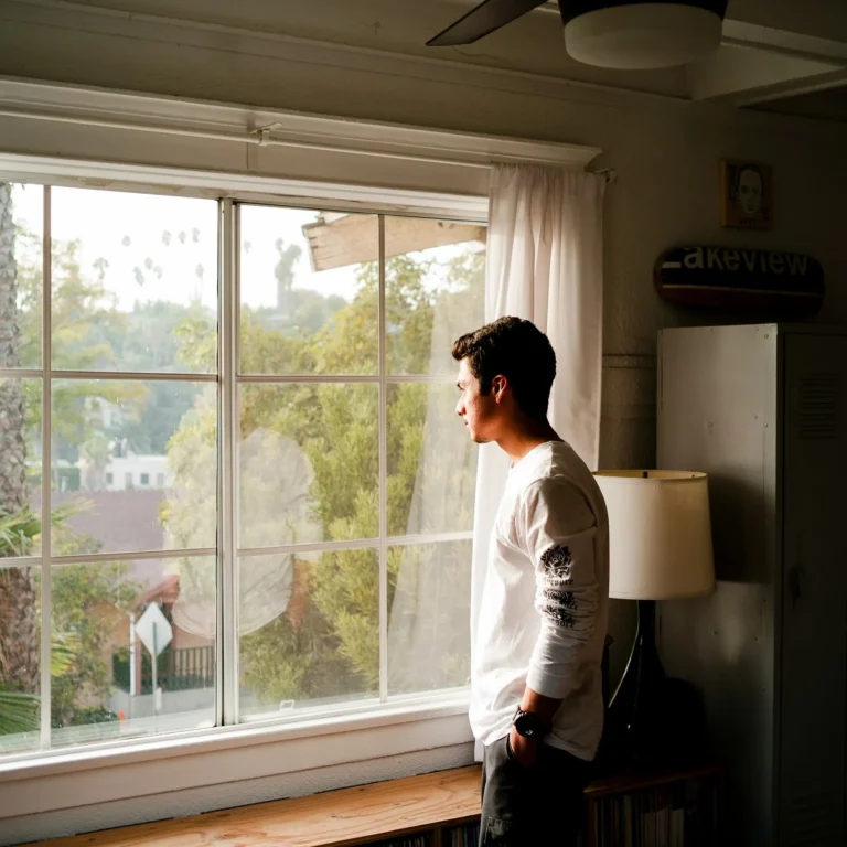 Young man standing at a bright window looking out, hands in pockets