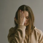 Woman in a beige hoodie standing with one hand covering half her face, eyes closed