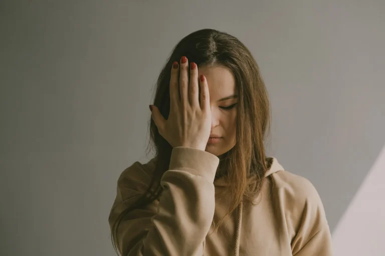 Woman in a beige hoodie standing with one hand covering half her face, eyes closed