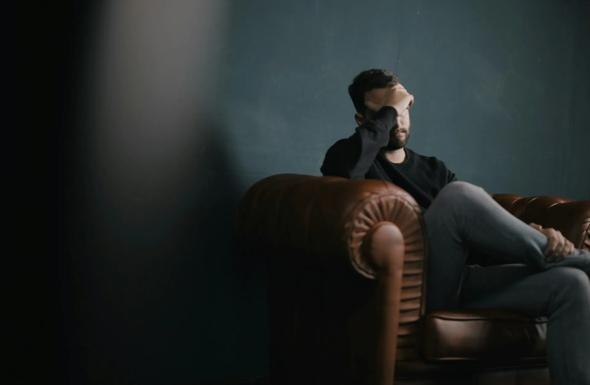 Man sitting alone on a leather sofa with his hand covering his face in a dim room