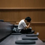 Young person in a white t-shirt sitting alone on auditorium steps with their head in their hands