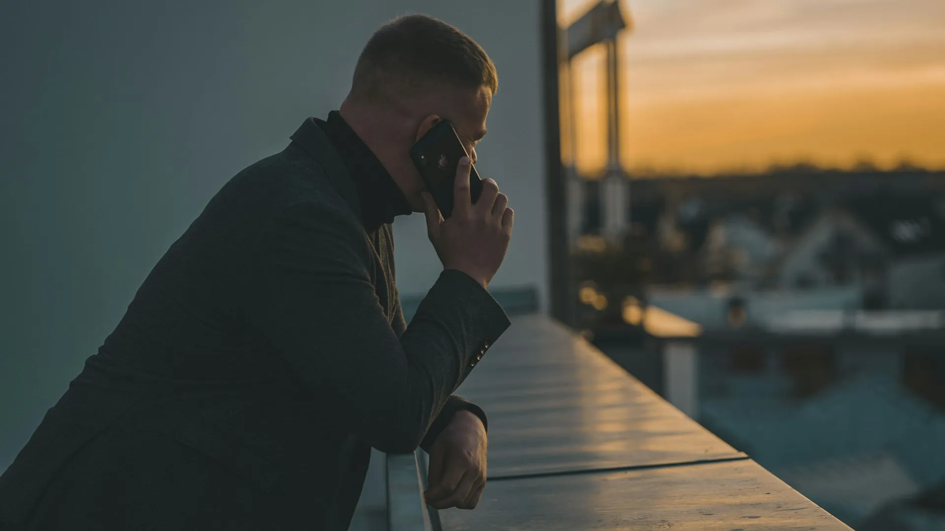 Man leaning on a railing making a phone call as the sun sets over rooftops