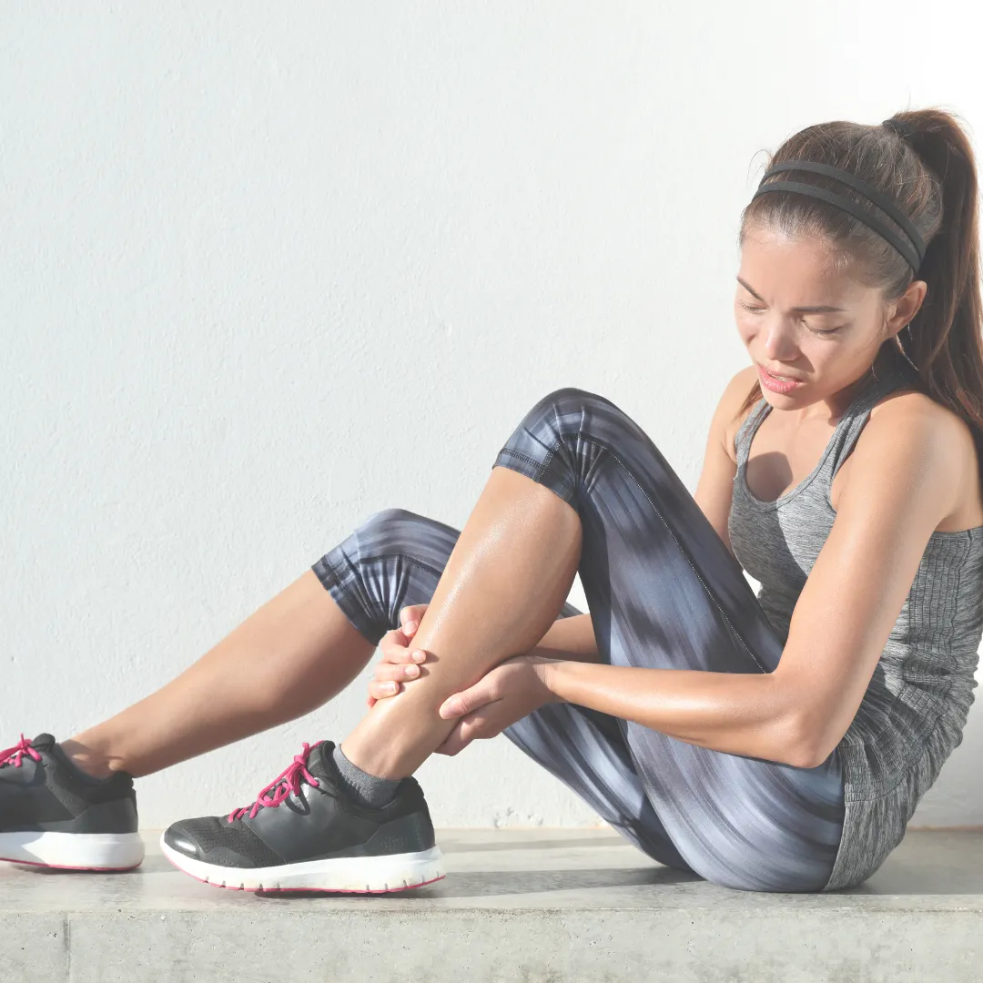 Female athlete in grey activewear holding her shin in pain due to delayed onset muscle soreness (DOMS) after a workout.
