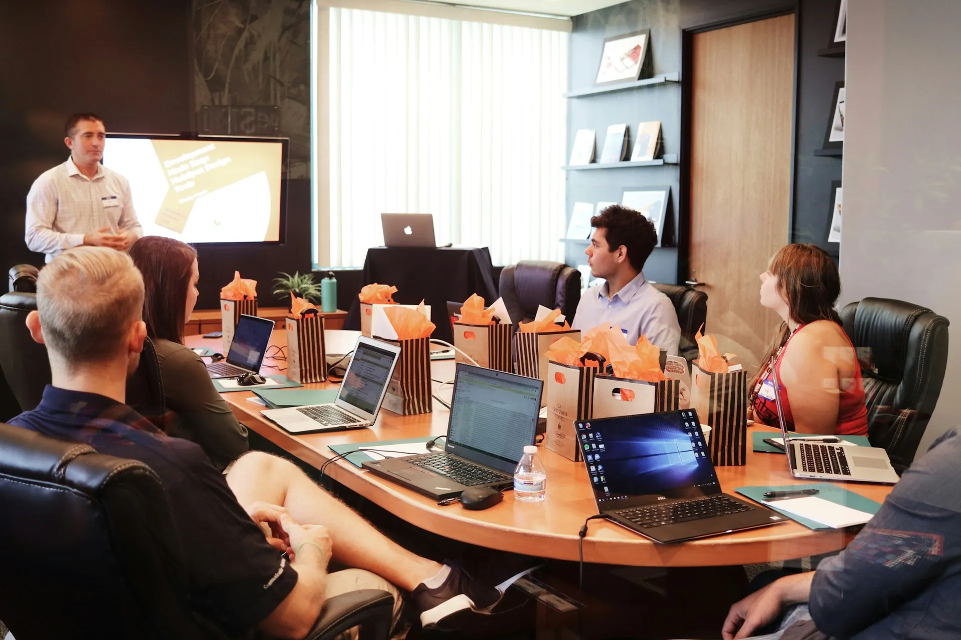 Man presenting to colleagues seated around a conference table with laptops and gift bags