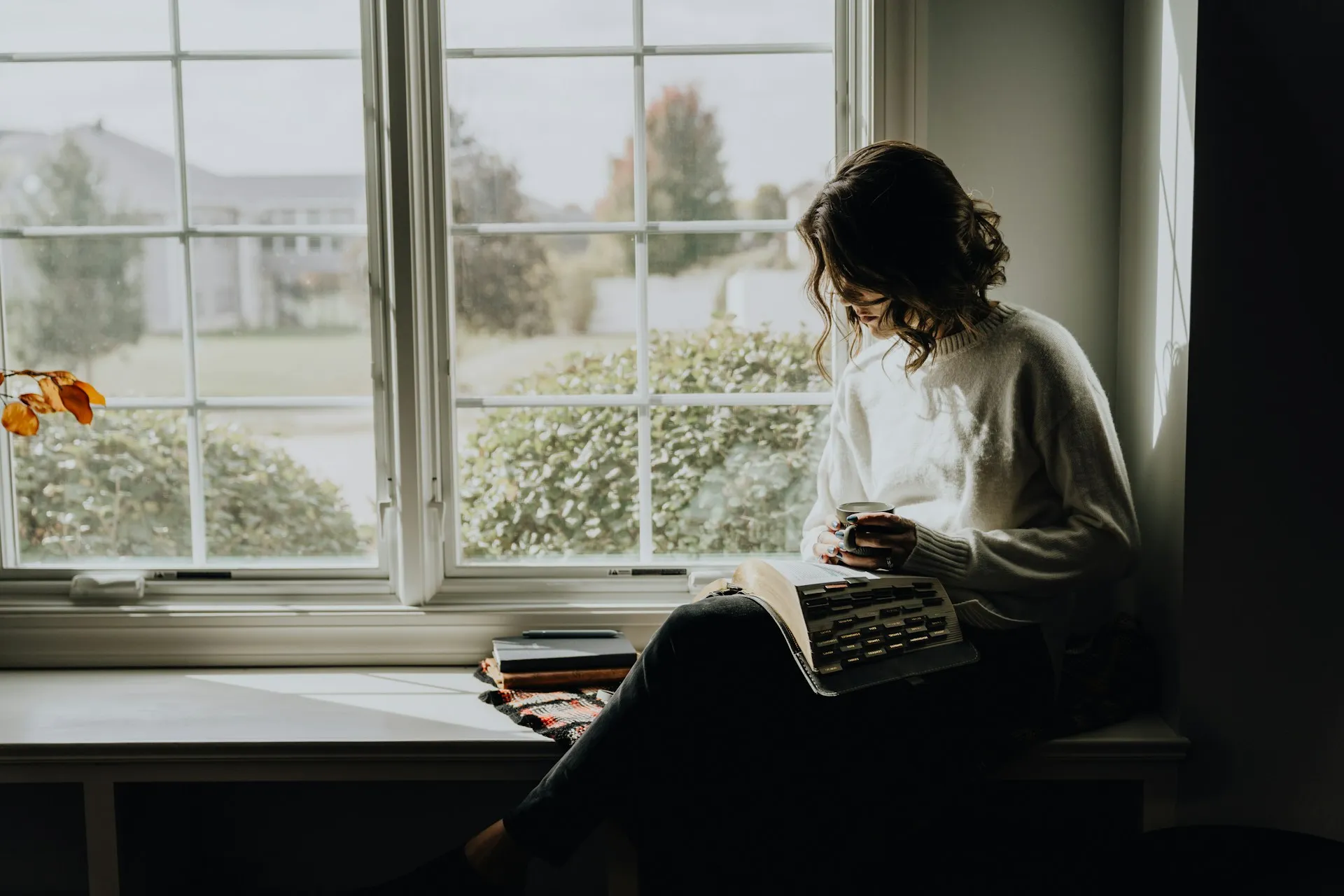 Woman sitting in a window seat reading a book and holding a mug in soft daylight