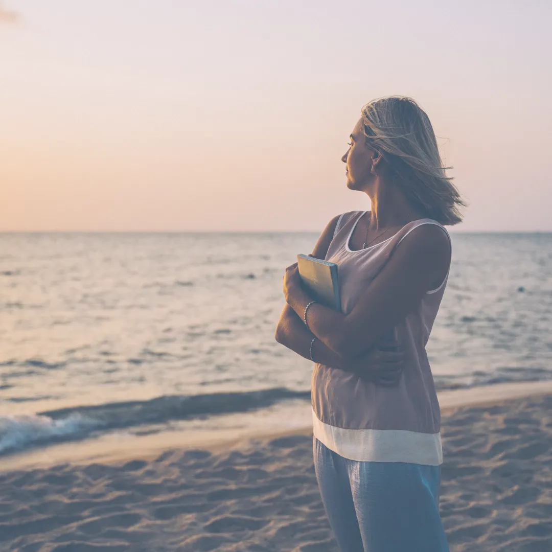 A person standing on a beach at sunset, holding a book to their chest and looking out at the ocean, symbolizing reflection and finding meaning in one's personal story.